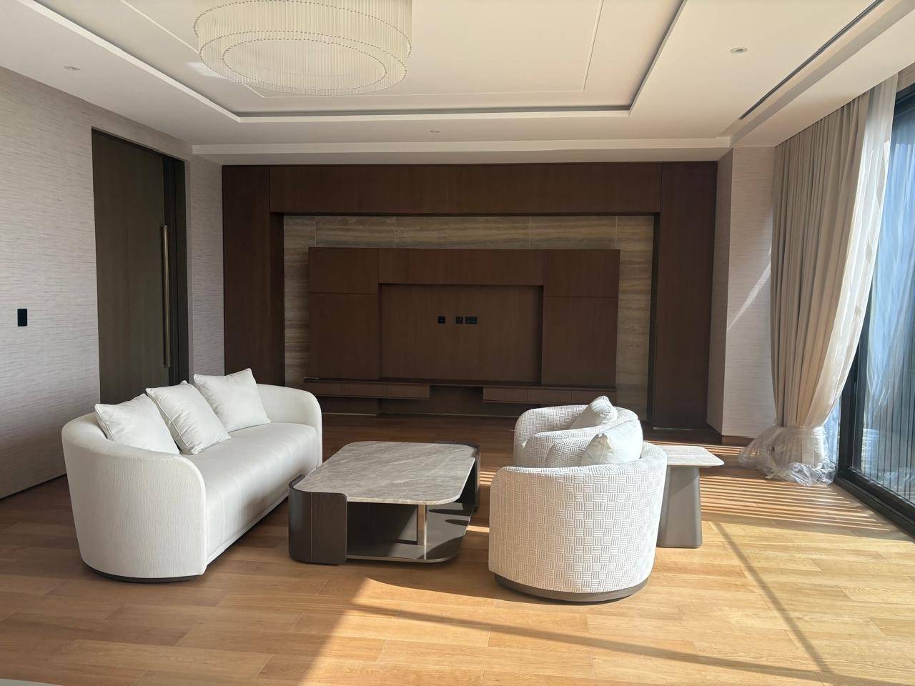 A bright modern living room in an Arabian Ranches villa with curved white sofa, patterned swivel armchairs, sleek marble coffee table, dark wood and stone accent wall, and contemporary chandelier