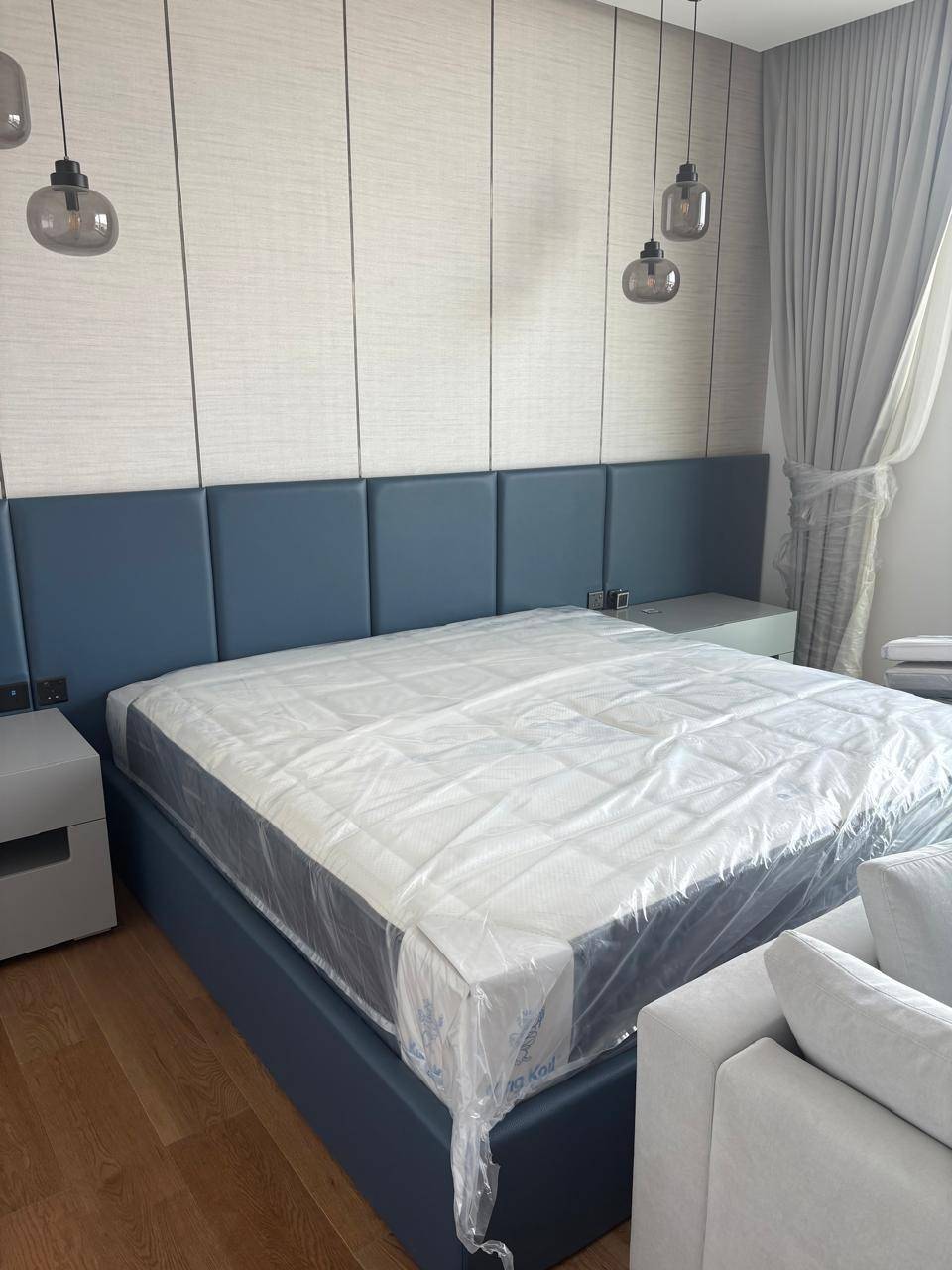 Freshly renovated modern bedroom with a blue upholstered bed, grey nightstands and smoked glass pendant lights against textured light grey wall paneling