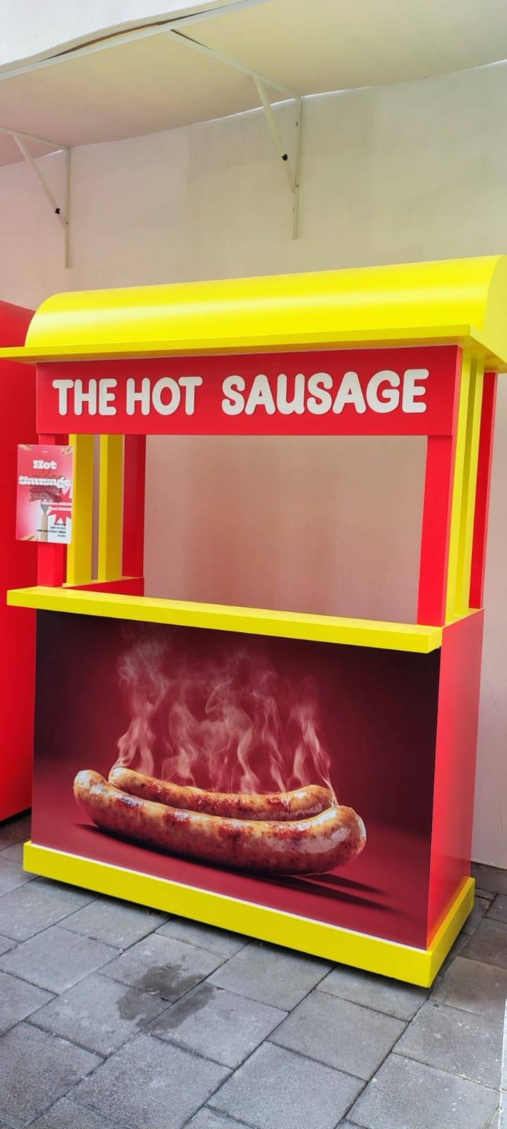 Custom fabricated red and yellow food stand with THE HOT SAUSAGE branding and a graphic of steaming grilled sausages, standing on grey paved flooring