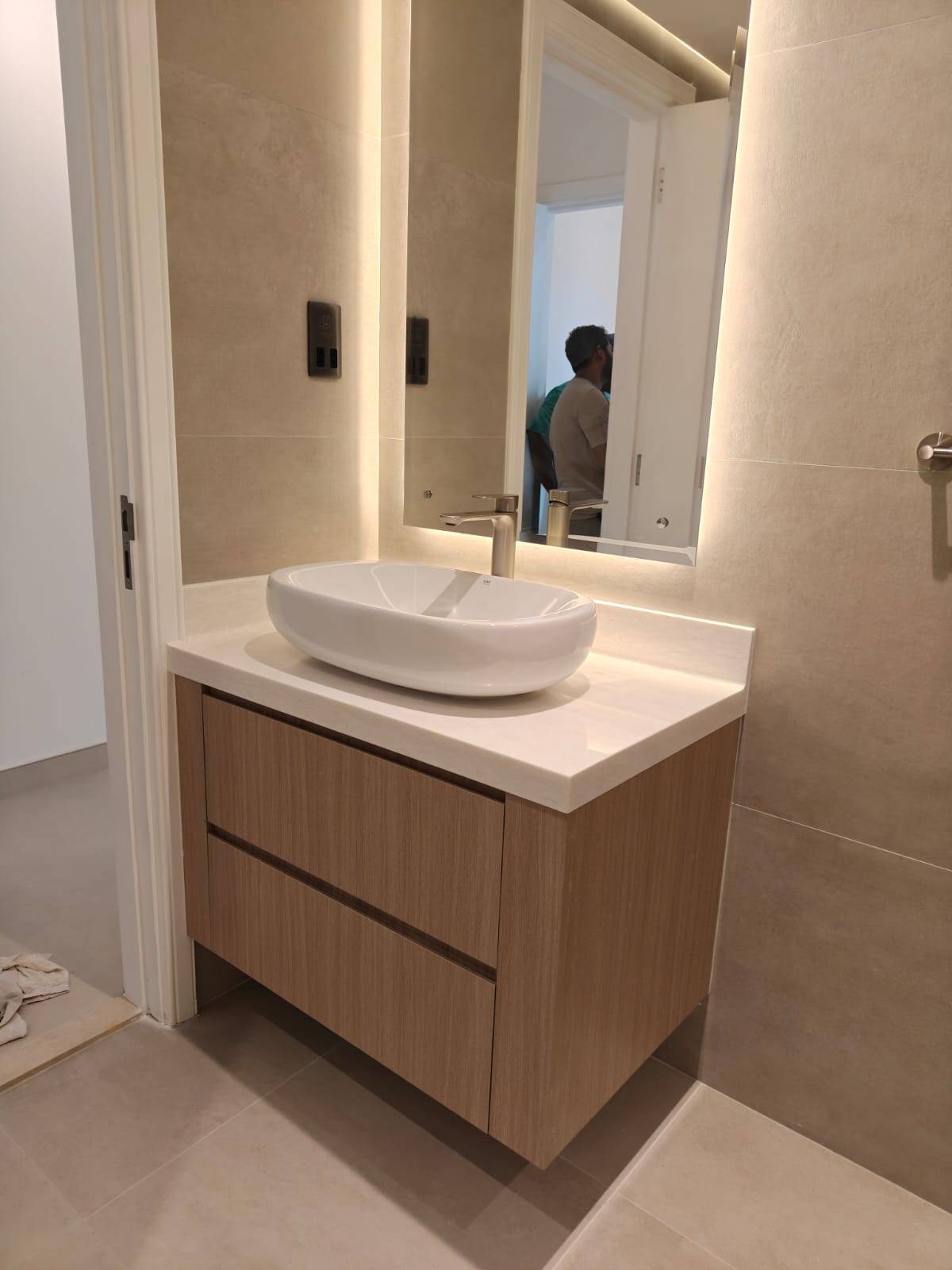 Freshly renovated modern bathroom in a Dubai villa extension with light wood floating vanity, white oval vessel sink, brushed nickel faucet, large mirror with integrated warm LED lighting and light beige tiled walls and floor