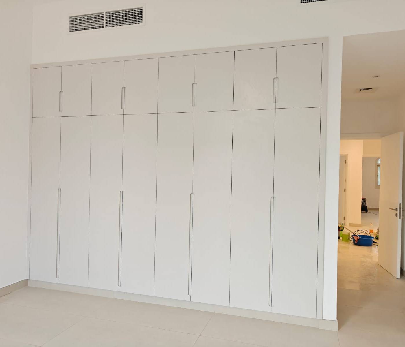 Freshly renovated room with a full wall light grey built in wardrobe with sleek integrated handles, white walls and light tiled flooring, with a glimpse into an adjacent hallway