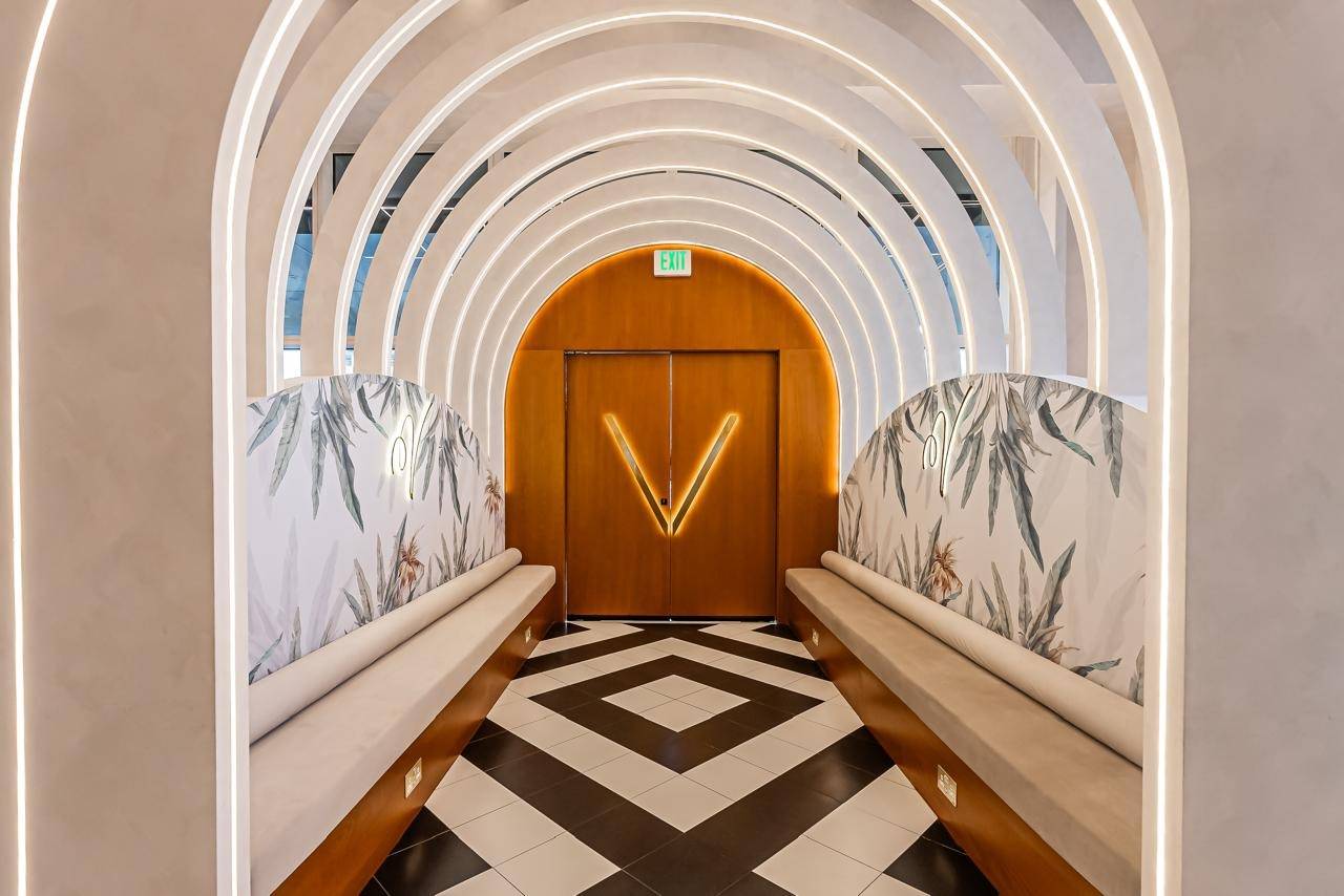 Interior hallway fit-out in Dubai with illuminated arches, a custom wooden door, built-in upholstered benches with botanical wallpaper backs and black and white geometric floor tiles