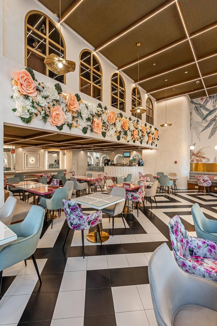Cafe interior in Dubai with a wall of large peach and white paper flowers, black and white geometric floor, arched windows and velvet seating under a gold ceiling with linear lighting