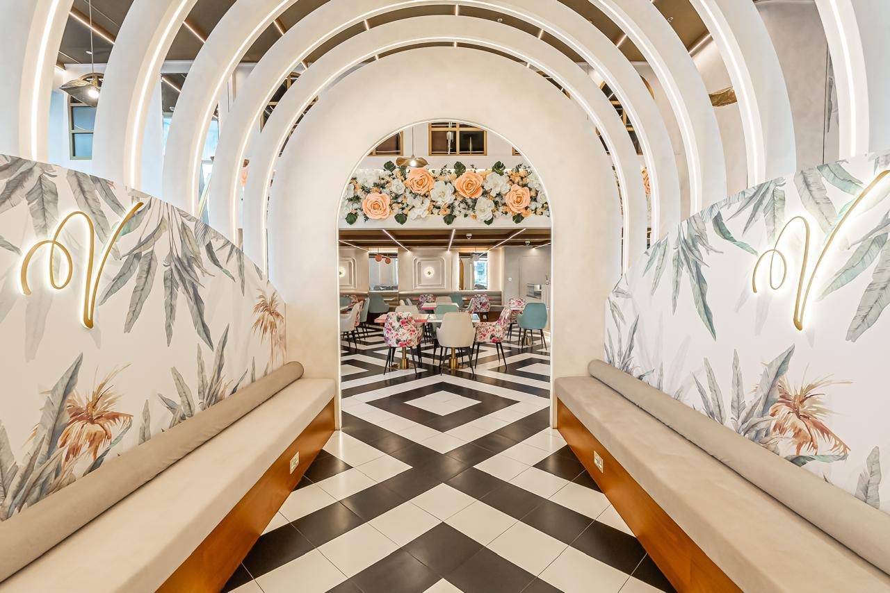 Cafe fit-out in Dubai with white arched passages, integrated lighting, a floral ceiling installation, geometric floor and banquette seating with tropical leaf wallpaper