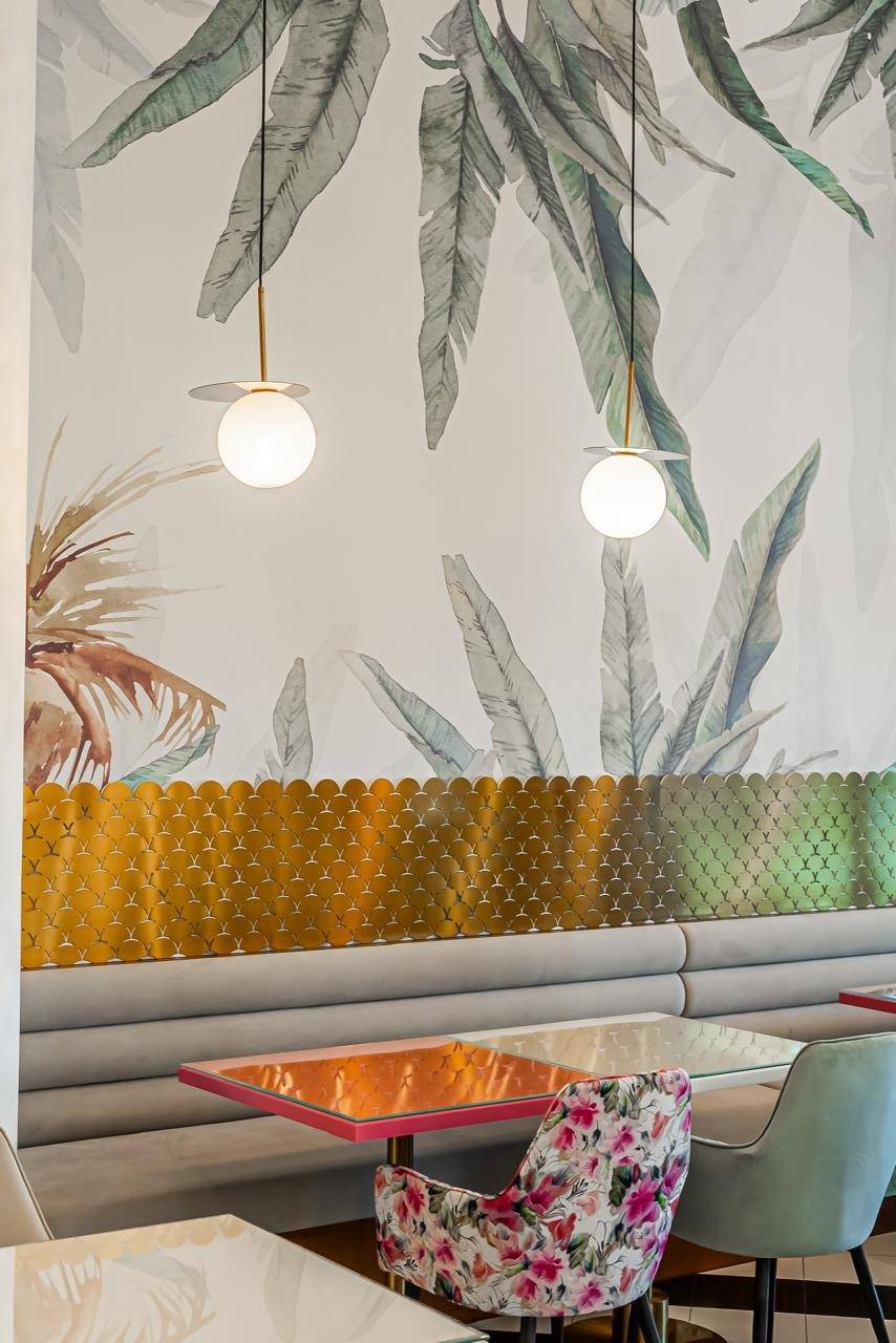Cafe interior fit-out in Dubai with tropical leaf wallpaper, a gold scale patterned screen, grey banquette seating and spherical pendant lights