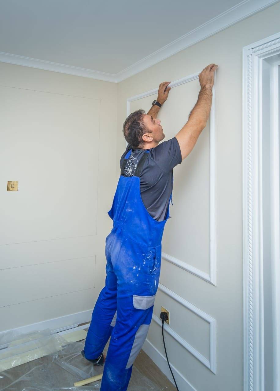 Worker in blue overalls installing white decorative wall moulding on a light beige wall during a Dubai apartment renovation
