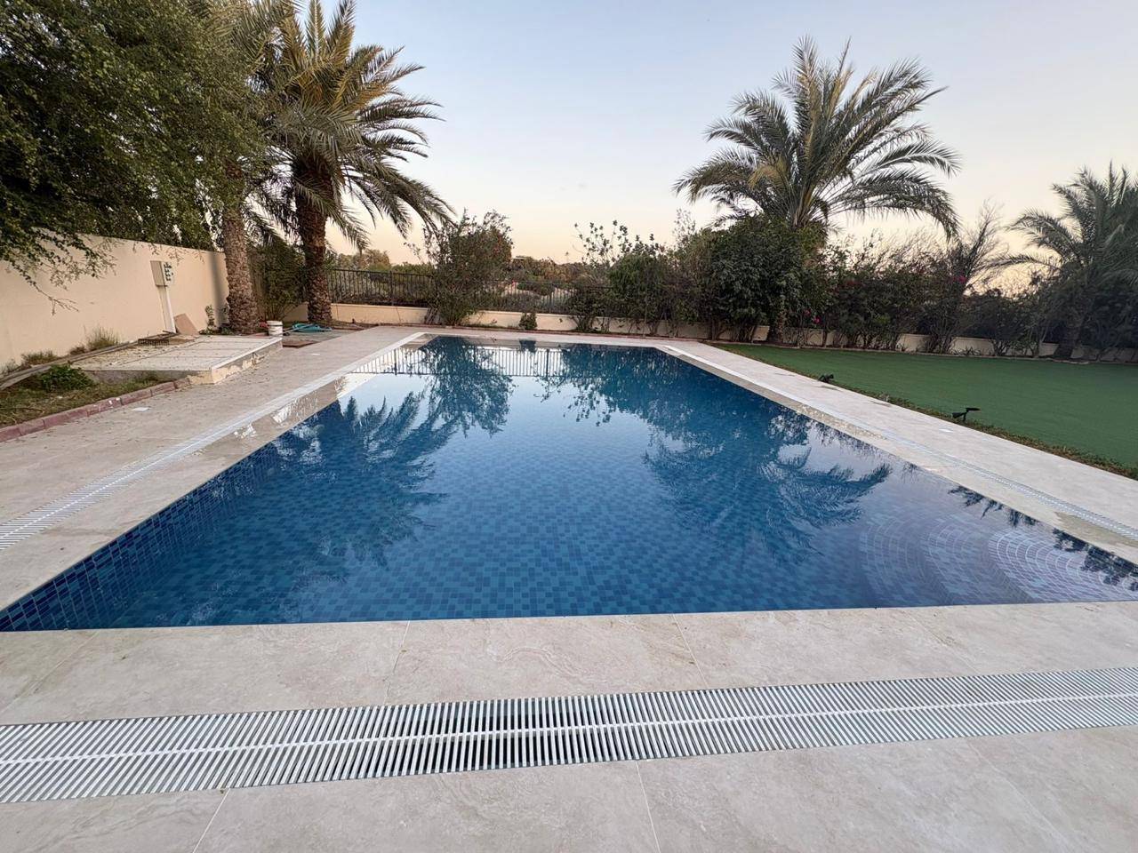 Newly finished Dubai villa pool with blue mosaic tiles, travertine coping and palm trees reflected in the water