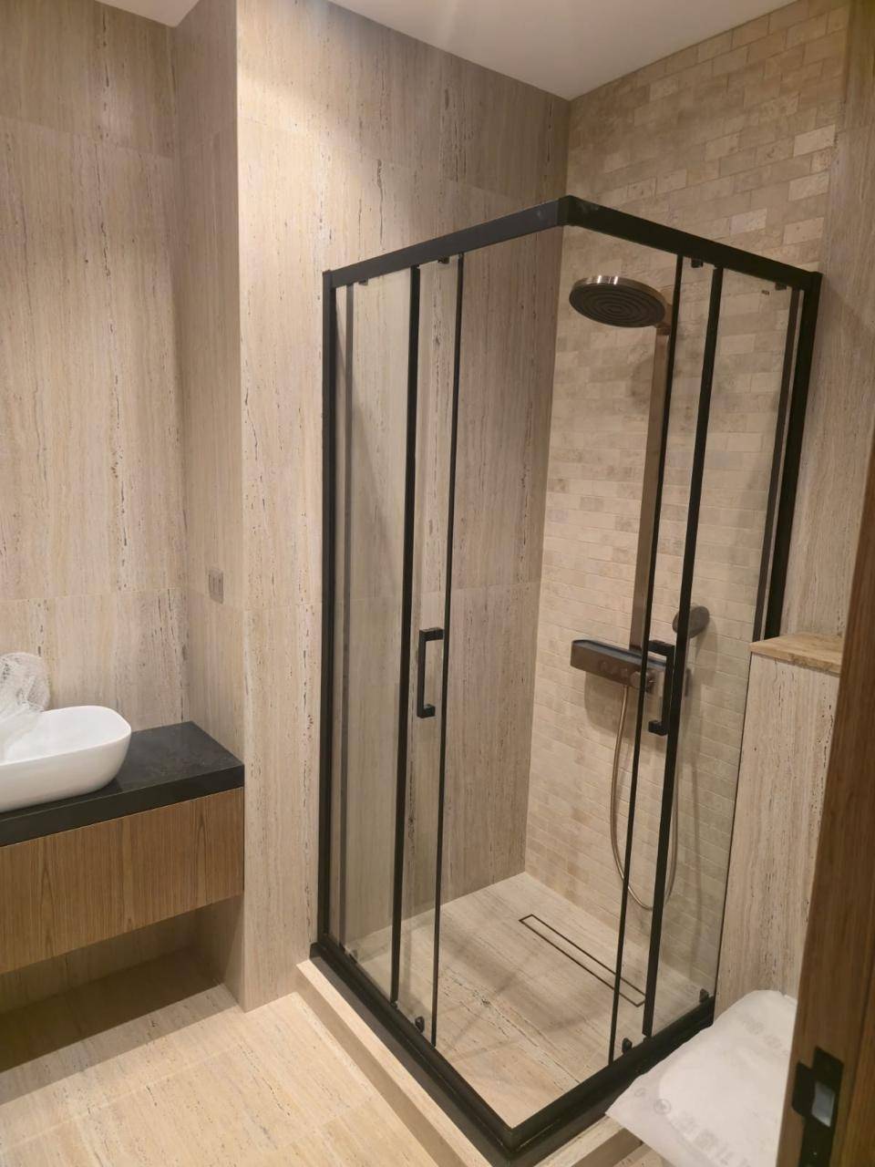 Renovated villa bathroom with a black framed glass shower, rain shower head and floating wooden vanity against beige natural stone tiles